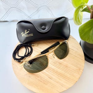 Peut inclure: Lunettes de soleil &agrave; monture en m&eacute;tal dor&eacute; avec verres vert fonc&eacute;, accompagn&eacute;es d'un &eacute;tui noir en similicuir portant le logo Ray-Ban Bausch & Lomb. Un cordon tress&eacute; noir avec des attaches en plastique transparent est inclus.