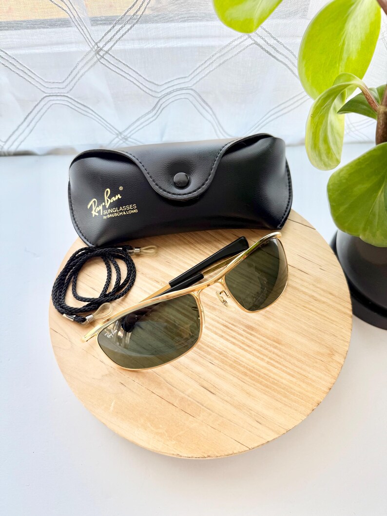 Peut inclure: Lunettes de soleil &agrave; monture en m&eacute;tal dor&eacute; avec verres vert fonc&eacute;, accompagn&eacute;es d'un &eacute;tui noir en similicuir portant le logo Ray-Ban Bausch & Lomb. Un cordon tress&eacute; noir avec des attaches en plastique transparent est inclus.