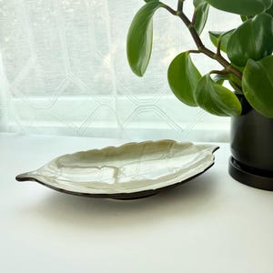 May include: A ceramic dish shaped like a leaf with a glossy, light green finish and dark brown edges. The surface features embossed vein details, mimicking the natural structure of a leaf. The dish is positioned horizontally on a flat surface.