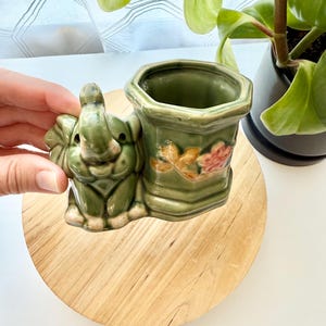 May include: A sage green ceramic planter featuring a sculpted elephant figure attached to the side of a hexagonal vessel. The vessel is decorated with a small, painted floral motif in shades of yellow and pink on its front exterior surface.