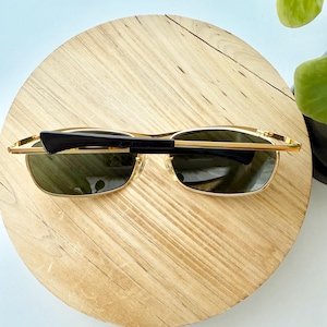 Peut inclure: Lunettes de soleil en m&eacute;tal dor&eacute; dot&eacute;es de verres rectangulaires teint&eacute;s vert fonc&eacute;. La monture pr&eacute;sente de fines branches en m&eacute;tal dor&eacute; et des embouts en plastique noir. Les lunettes sont pli&eacute;es, montrant la face avant et une branche.