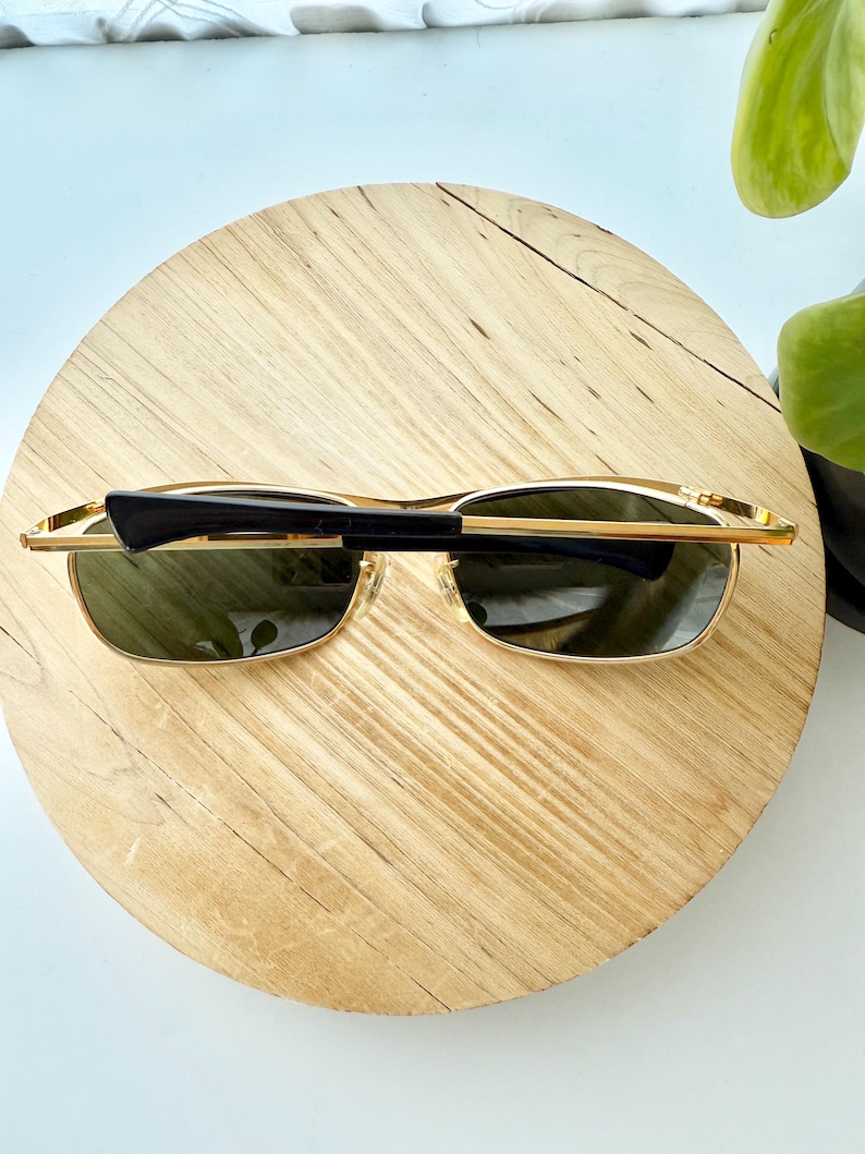 Peut inclure: Lunettes de soleil en m&eacute;tal dor&eacute; dot&eacute;es de verres rectangulaires teint&eacute;s vert fonc&eacute;. La monture pr&eacute;sente de fines branches en m&eacute;tal dor&eacute; et des embouts en plastique noir. Les lunettes sont pli&eacute;es, montrant la face avant et une branche.