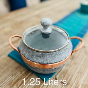 K&ouml;nnte beinhalten: Ein grauer Steintopf mit Kupferrand und Glasdeckel. Der Topf hat einen Holzgriff auf dem Deckel. Der Topf steht auf einem blauen und gr&uuml;nen Untersetzer auf einem Holztisch. 1,25 Liter.