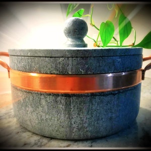 May include: A round, gray stone pot with a copper band around the rim and a copper handle on each side. The pot has a lid with a round knob on top. A green plant is growing out of the pot.