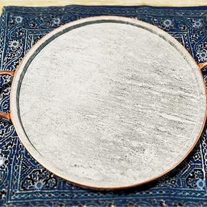 May include: A round, gray stone serving tray with copper-colored handles. The tray sits on a blue patterned cloth with a floral design. The tray is ideal for serving appetizers or desserts.