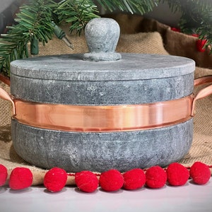 May include: A gray soapstone pot with a copper band around the rim and a copper handle. The pot has a lid with a knob on top. The pot is sitting on a burlap surface with red pom-poms.