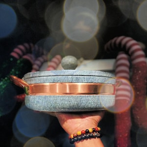 May include: A round, blue-grey cooking pot with a copper band and handle. The pot has a matching stone lid with a round knob. The pot is being held up by a person. The background is blurred with bokeh lights.