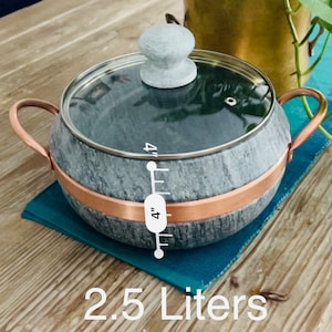 K&ouml;nnte beinhalten: Ein grauer Specksteintopf mit Kupferrand und Glasdeckel. Der Topf hat einen Holzgriff und ein Lineal an der Seite, das "4 Zoll" anzeigt. Der Topf steht auf einem blauen Untersetzer und einem Holztisch. Der Text "2,5 Liter" ist unter dem Topf aufgedruckt.