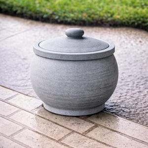 May include: A gray stone garden pot with a lid. The pot has a round body and a flat base. The lid has a small, round handle. The pot is sitting on a brick patio.