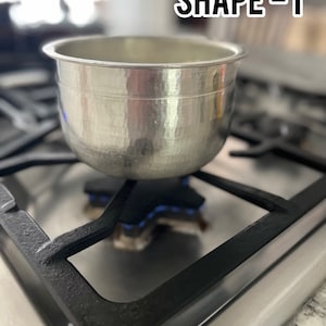 May include: A silver hammered metal pot sits on a black gas stovetop burner with a blue flame. The text "SHAPE - 1" is visible in the upper right corner of the image.