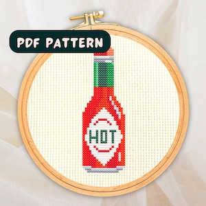 Op de afbeelding: Een kruissteekpatroon van een rode hete sausfles met een groene dop en het woord "HOT" in het wit. Het ontwerp is geplaatst in een houten borduurring. De tekst "PDF PATTERN" staat in een donkergroene rechthoek bovenaan.
