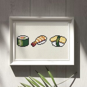 May include: A white framed cross-stitch embroidery featuring three different types of sushi: a green maki roll, a shrimp nigiri, and a yellow tamago nigiri.