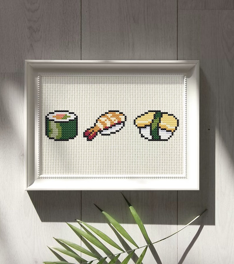 May include: A white framed cross-stitch embroidery featuring three different types of sushi: a green maki roll, a shrimp nigiri, and a yellow tamago nigiri.