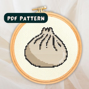 May include: A cross-stitch pattern of a white and beige steamed bun with pleats. The pattern is in a wooden embroidery hoop.