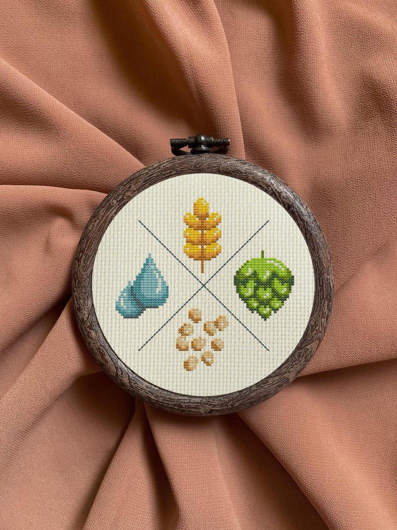 May include: A round embroidery hoop with a white fabric background featuring a cross-stitch design of four elements: a blue water drop, a yellow wheat stalk, a green hop cone, and a cluster of beige barley grains.