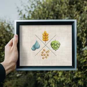 May include: A framed cross-stitch design featuring four elements: a blue water drop, a green hop, a yellow wheat stalk, and a cluster of brown barley. The design is on a beige linen background.