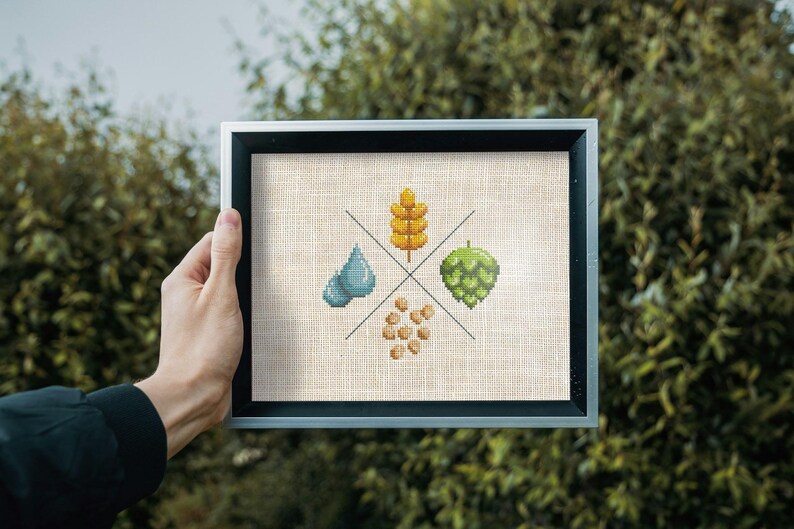 May include: A framed cross-stitch design featuring four elements: a blue water drop, a green hop, a yellow wheat stalk, and a cluster of brown barley. The design is on a beige linen background.
