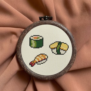 May include: A close-up of a round embroidery hoop with a white fabric background. The fabric is embroidered with three different types of sushi: a maki roll, a nigiri with a shrimp topping, and a nigiri with a yellow topping. The embroidery is done in a pixelated style.