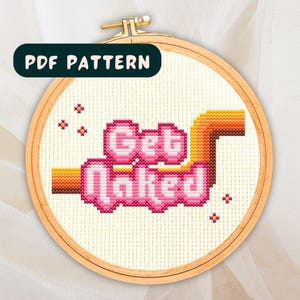 May include: A cross-stitch pattern featuring the phrase "Get Naked" in pink letters with a yellow and orange background. The pattern is on a white background and is framed in a wooden hoop.