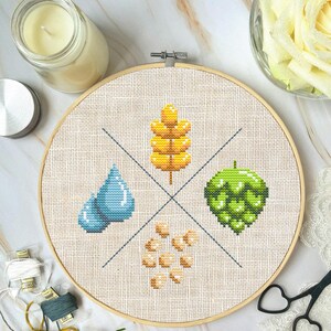 May include: A cross-stitch embroidery hoop featuring a design of four elements: a blue water drop, a green hop, a yellow wheat stalk, and a cluster of beige barley. The hoop is on a white marble surface.