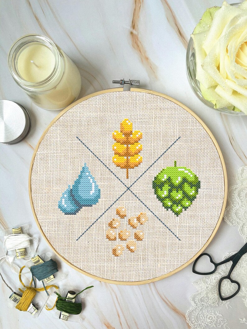 May include: A cross-stitch embroidery hoop featuring a design of four elements: a blue water drop, a green hop, a yellow wheat stalk, and a cluster of beige barley. The hoop is on a white marble surface.