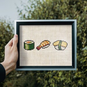 May include: A framed cross-stitch piece depicting three different types of sushi: a green maki roll, a shrimp nigiri, and a yellow tamago nigiri.