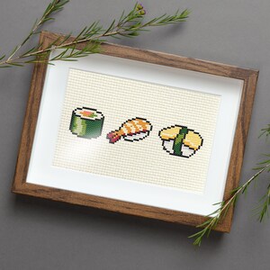 May include: A framed cross-stitch embroidery depicting three different types of sushi: a maki roll, a nigiri with shrimp, and a temaki hand roll. The embroidery is done in a pixelated style, with bright colours and a simple design.