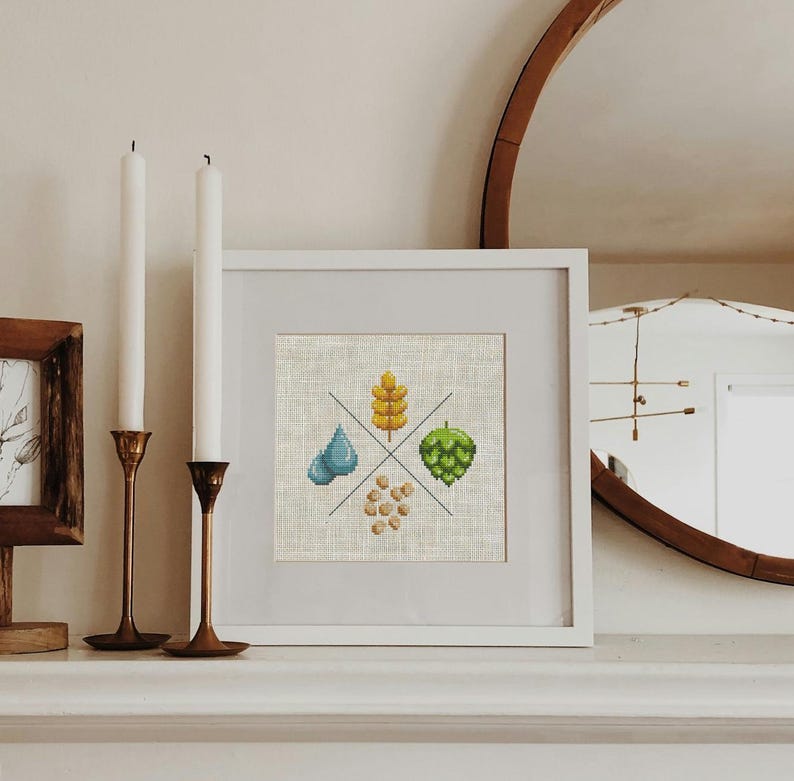 May include: A white framed cross-stitch embroidery featuring four elements: a blue water drop, a yellow wheat stalk, a green hop cone, and a pile of brown barley. The embroidery is displayed on a white mantelpiece with two candlesticks and a framed print.