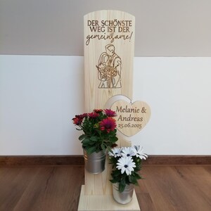 Könnte beinhalten: Holzhochzeitsschild mit dem Text "DER SCHÖNSTE WEG IST DER gemeinsame!" und einer Paarillustration. Es hat zwei Metalleimer mit Blumen und eine herzförmige Plakette mit Namen und Datum.