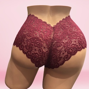 May include: Burgundy lace underwear with a high waist design. The intricate floral lace pattern covers the front and back of the garment. The underwear is displayed on a mannequin against a pink background.