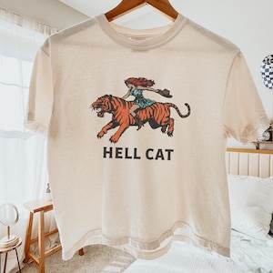 Könnte beinhalten: Ein beiges T-Shirt mit einem Aufdruck einer Frau, die auf einem Tiger reitet. Unter dem Aufdruck steht der Text "HELL CAT".