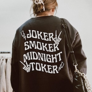 May include: Black sweatshirt with white text that reads "JOKER SMOKER MIDNIGHT TOKER". The design includes lightning bolts and skeleton hands. The sweatshirt is part of a clothing collection.