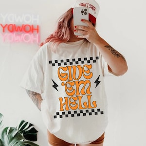 Pode incluir: Camiseta branca com uma borda xadrez preta e branca e o texto "Give 'em Hell" em laranja. O texto está em um estilo de fonte retrô.
