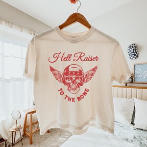 May include: A cream colored cropped t-shirt with a red graphic of a skull wearing a helmet and aviator sunglasses with wings. The text "Hell Raiser to the Bone" is printed in red below the skull.