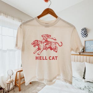 Könnte beinhalten: Ein cremefarbenes T-Shirt mit einer roten Grafik einer Frau, die auf einem Tiger reitet. Der Text "HELL CAT" ist in Rot unter der Grafik gedruckt.