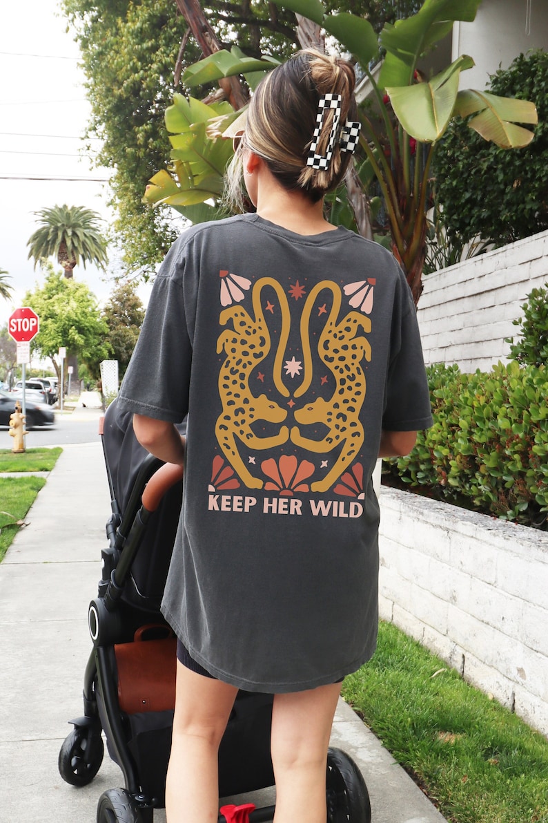 May include: A charcoal grey t-shirt with a graphic of two stylized leopards facing each other. The graphic is in shades of orange, yellow, and pink. The text "KEEP HER WILD" is printed below the graphic.
