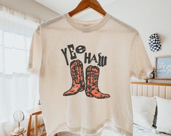 May include: A cream coloured t-shirt with short sleeves and a cropped hem. The shirt has a graphic print of two cowboy boots and the text "Yee Haw" in black.