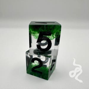Puede incluir: Dos dados de resina transparente con números negros e inclusiones verdes. El dado superior muestra un gran "5" negro, mientras que el dado inferior muestra un "2" negro. Los dados están apilados, mostrando un diseño único.