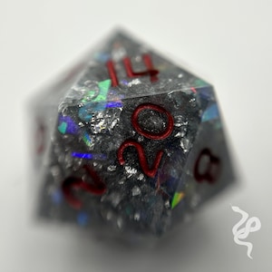 May include: A clear, iridescent, resin d20 dice with silver glitter and red numbers. The numbers 14 and 20 are visible.