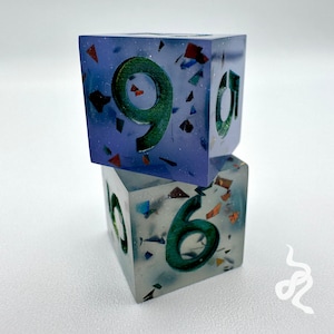 May include: Two dice with a glittery finish. The top die is purple with a green number nine. The bottom die is clear with a green number six.