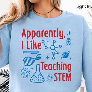 May include: Light blue sweatshirt with the text "Apparently, I Like Teaching STEM" in red and blue. The design includes science-related graphics such as a DNA strand, a rocket, and a molecule.