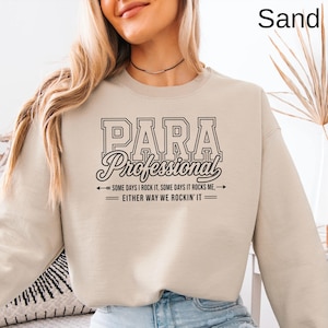 May include: A sand-colored crewneck sweatshirt featuring black text that reads PARA PROFESSIONAL, followed by the phrase SOME DAYS I ROCK IT, SOME DAYS IT ROCKS ME, EITHER WAY WE ROCKIN' IT, accented with arrows and horizontal lines.