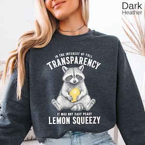 May include: A dark heather gray crewneck sweatshirt featuring a graphic of a raccoon holding a yellow lemon. The text reads: IN THE INTEREST OF FULL TRANSPARENCY IT WAS NOT EASY PEASY LEMON SQUEEZY. The design is printed in white and yellow ink.