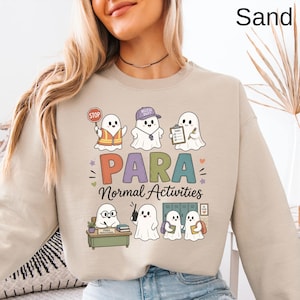May include: A sand-colored crewneck sweatshirt featuring a graphic of six cartoon ghosts. The text reads "PARA Normal Activities". The ghosts are depicted in various school-related roles, including holding a stop sign, wearing a cap, and using a walkie-talkie, with a desk and lockers in the background.