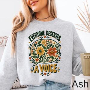 May include: A light gray crewneck sweatshirt featuring a central graphic of a speech bubble filled with various flowers in shades of yellow, orange, and green. The text "EVERYONE DESERVES A VOICE" is printed in dark green and gold lettering.