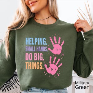 May include: A military green crewneck sweatshirt featuring the text HELPING. SMALL HANDS. DO BIG. THINGS. in light blue, purple, pink, and orange, accompanied by two pink handprint graphics on the right side of the chest area.