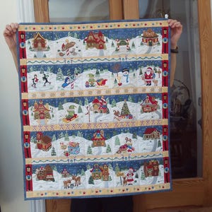 May include: A rectangular Christmas quilt with multiple rows of winter scenes. Each row features houses, snow-covered landscapes, and festive characters like Santa Claus and snowmen. The quilt has a blue border and is held up by a person.