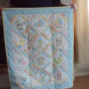 May include: A baby quilt with a light blue border and a yellow trim. The quilt features embroidered images of a sheep, duck, sailboat, teddy bear, and other baby-themed designs in pastel colours. The quilt is held up by a person.