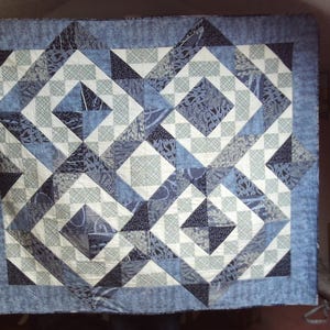 May include: A handmade quilt with a geometric pattern in shades of blue, white, and grey. The design features squares, triangles, and diamond shapes, creating a visually appealing patchwork. The quilt has a light blue border.