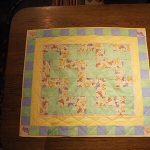 May include: A square baby quilt with a patchwork design. The centre features a pattern of green and patterned fabric squares. The quilt is bordered by yellow, pink, and blue squares. The quilt is likely handmade.